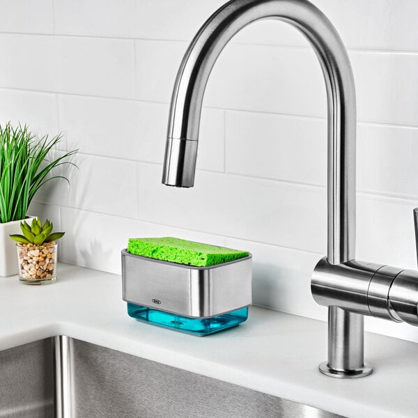 An OXO Good Grips soap dispensing sponge holder with a green sponge on the counter above a stainless steel sink.