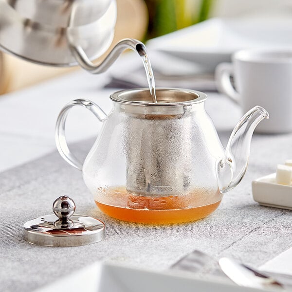 A glass teapot with a metal tea strainer being used to pour tea.