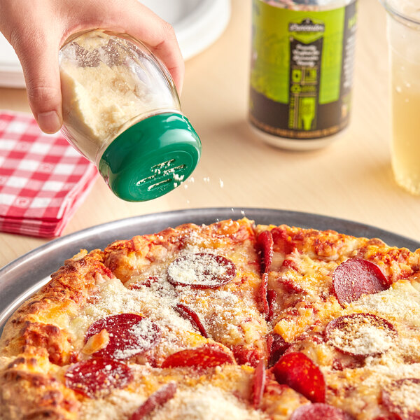 A hand holding a Tablecraft glass swirl shaker with a green slotted top pouring seasoning onto a pepperoni pizza.