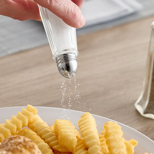 A hand pouring salt from a Tablecraft salt shaker onto a plate of french fries.