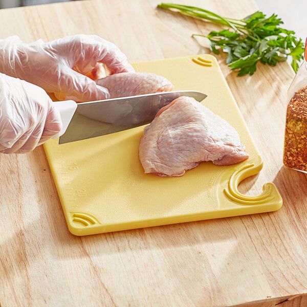 A person in gloves using a knife to cut meat on a yellow San Jamar cutting board.