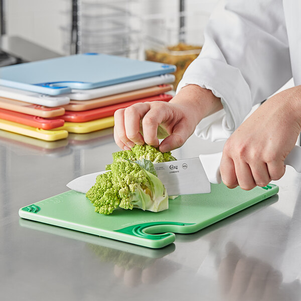 A person in a white coat cutting a piece of broccoli on a blue and white San Jamar Saf-T-Grip cutting board.