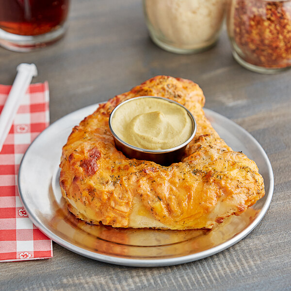 A pizza on a wide rim pizza pan with a bowl of white sauce on a table with a red and white checkered tablecloth.