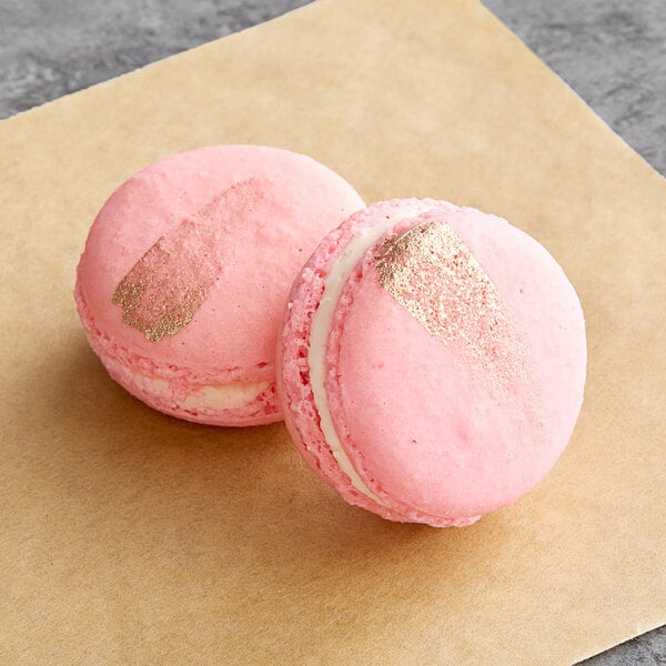 Two pink Macaron Centrale macarons with gold foil on them on a brown surface.