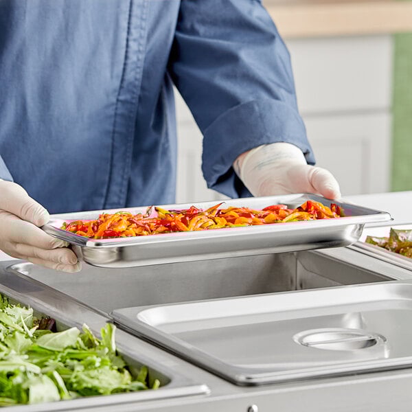 A person in a blue uniform holding a Vollrath stainless steel steam table pan filled with food.