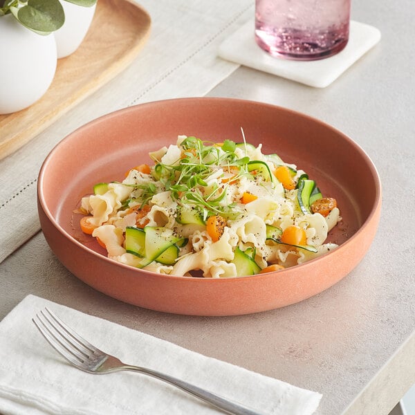 A Acopa Terra Cotta porcelain pasta bowl filled with pasta, vegetables, and greens with a fork on a white table.