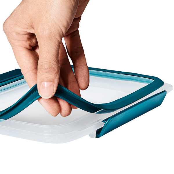A hand holding a white OXO Prep & Go rectangular food storage container with a blue lid.