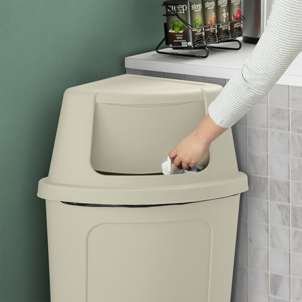A woman opens a Lavex beige corner round trash can lid in a kitchen.