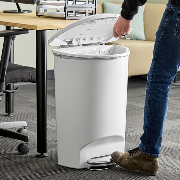 A person in jeans using a simplehuman white semi-round step-on trash can to throw away paper.