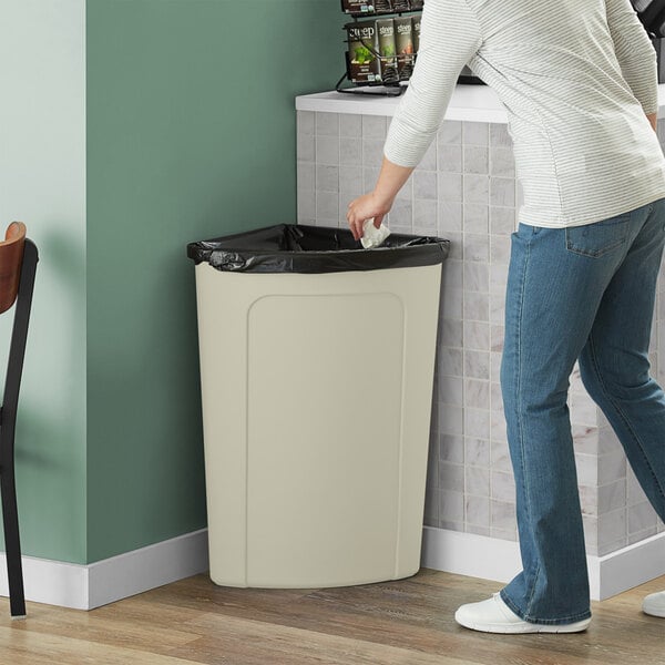 A person in jeans puts a bag in a beige Lavex corner round trash can.