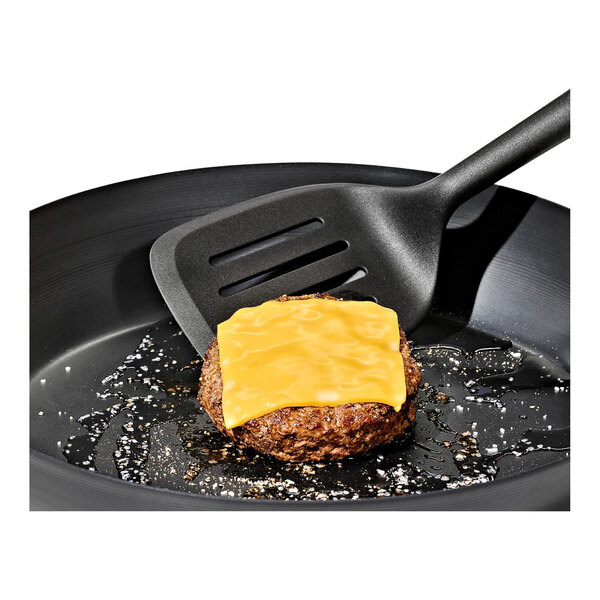 A black nylon perforated turner spatula being used to cook a cheeseburger in a frying pan.