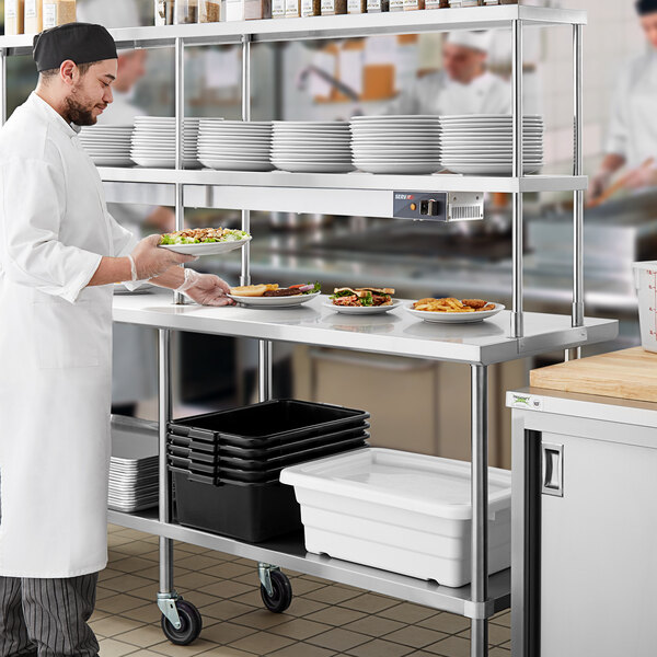 A man in a white coat standing at a stainless steel Regency work table holding a plate of food.