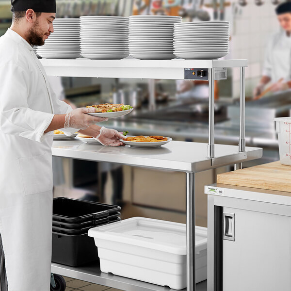 A chef in a white coat standing at a Regency work table holding plates of food.