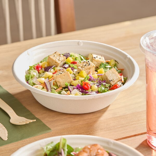 A white compostable fiber bowl filled with a colorful salad and tofu cubes, placed on a wooden table.