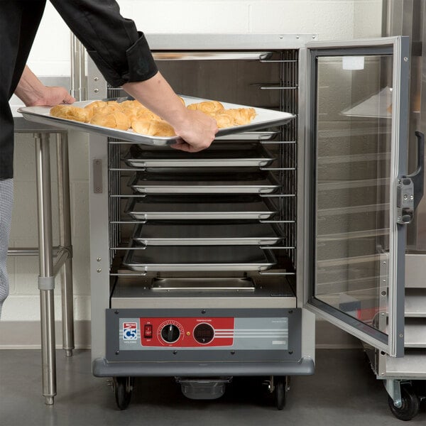 A person holding a tray of pastries in a Metro half-size holding cabinet with a clear door.
