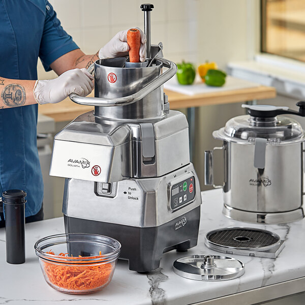A person using an AvaMix commercial food processor to shred carrots in a stainless steel bowl.