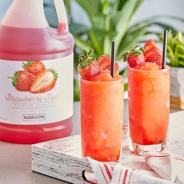 A couple of glasses of red Narvon Strawberry Slushy with straws sitting next to a pitcher of strawberry juice.
