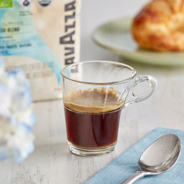 A glass cup of Lavazza decaf espresso with a spoon on a table with a bag of Lavazza coffee.