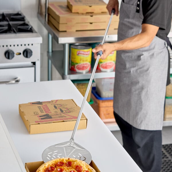 A stainless steel round perforated pizza peel with a long handle being used in a commercial kitchen.