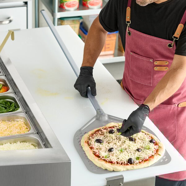 A square perforated aluminum pizza peel with a long handle being used to prepare a pizza.