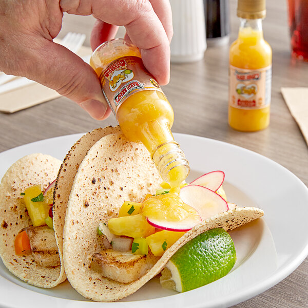 A hand pouring Marie Sharp's Orange Pulp Habanero Hot Sauce onto a taco.