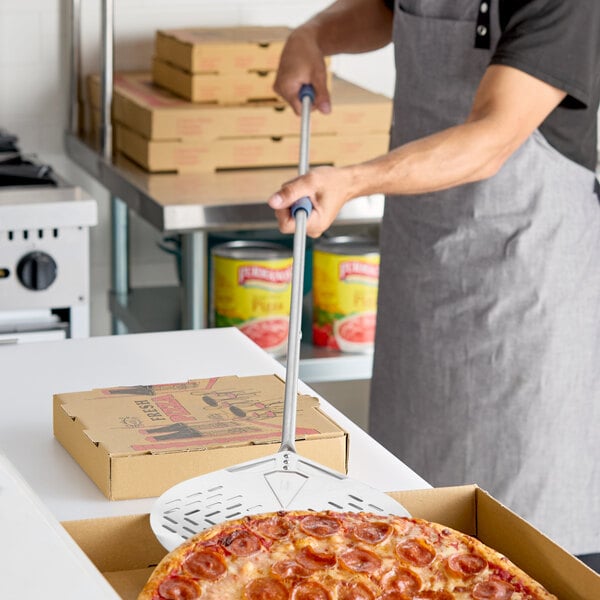 A stainless steel round perforated pizza peel with a long handle being used to place a pepperoni pizza into a box.