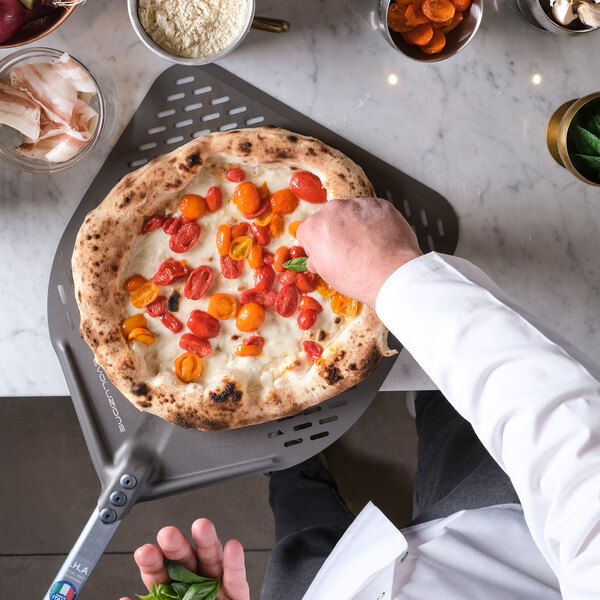 A person in a white coat is garnishing a freshly baked pizza with basil leaves on a perforated pizza peel, surrounded by various ingredients on a marble countertop.
