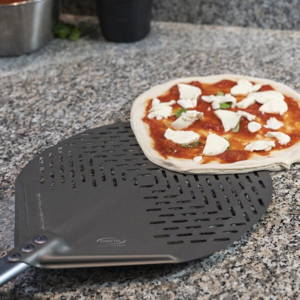 A round, perforated metal pizza peel with a partially prepared pizza resting on it, placed on a granite countertop.