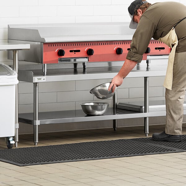 A man wearing an apron and holding a silver pot standing at a Regency stainless steel equipment stand in a school kitchen.