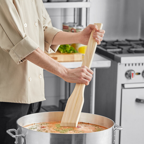 A person using a Choice 24" Wood Paddle to stir a pot of soup.