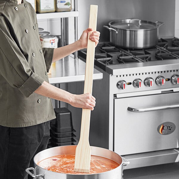 A person holding a Choice wood paddle over a large pot of soup.