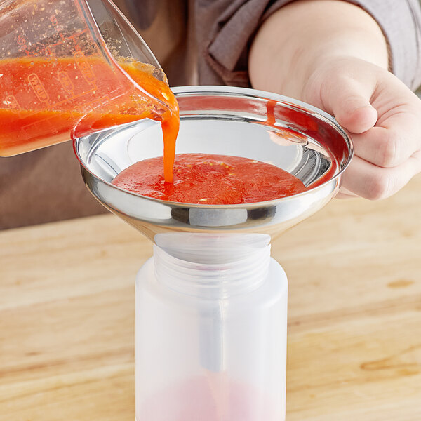 A person pouring red liquid into a stainless steel funnel with a strainer.