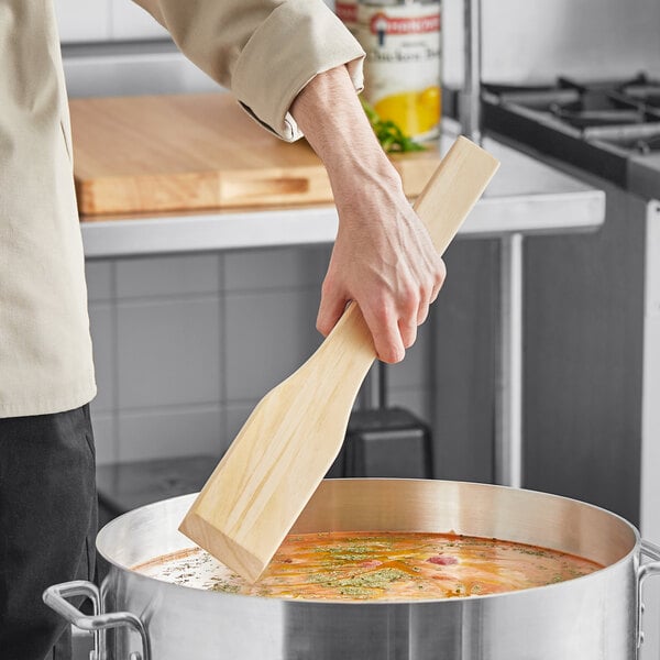 A hand holding a Choice 18" wood paddle over a pot of soup.