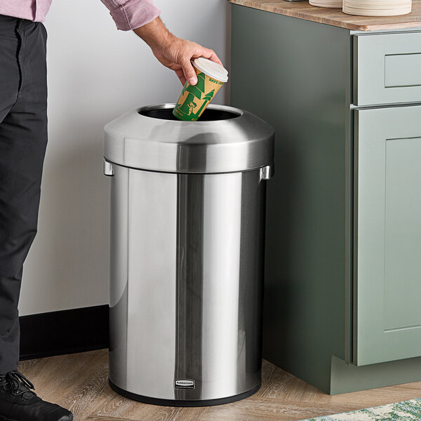 A person standing in a home kitchen places a coffee cup in a Rubbermaid stainless steel trash can.