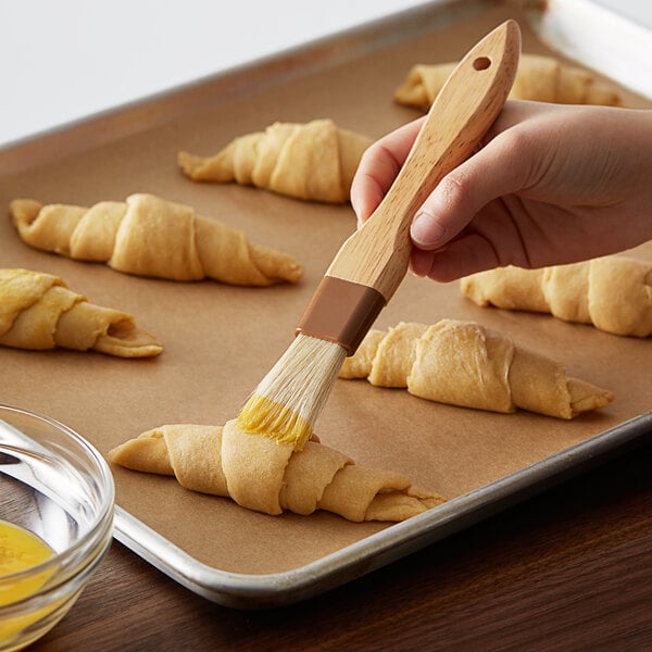 A person using a 1" boar bristle pastry brush to baste croissants.