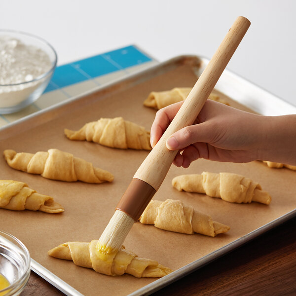 A hand holding a 1"W round boar bristle pastry brush with a wooden handle over croissants.