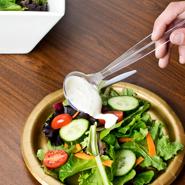 A person using a Fineline clear plastic ladle to pour dressing on a salad.