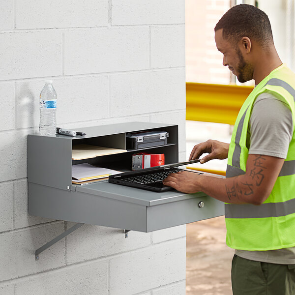 A man in a safety vest using a Lavex wall mount shop desk with a laptop.