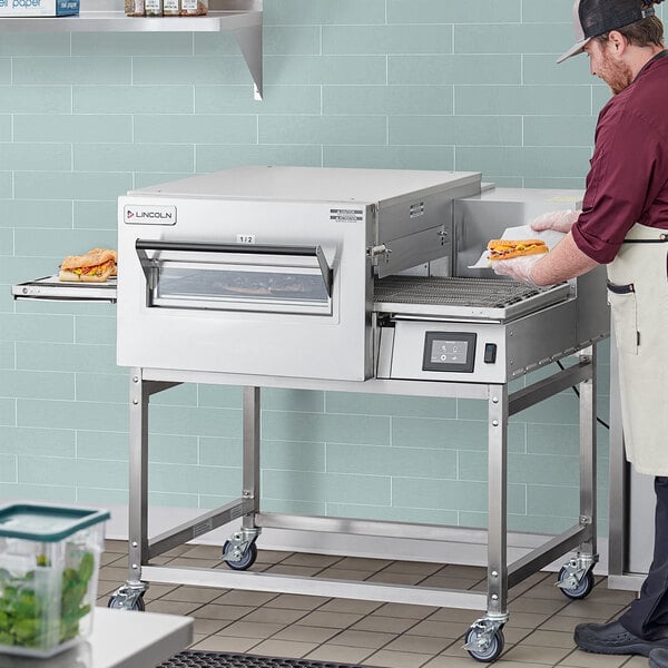 A man using a Lincoln Impinger II liquid propane conveyor oven with a glass door to cook a pizza.