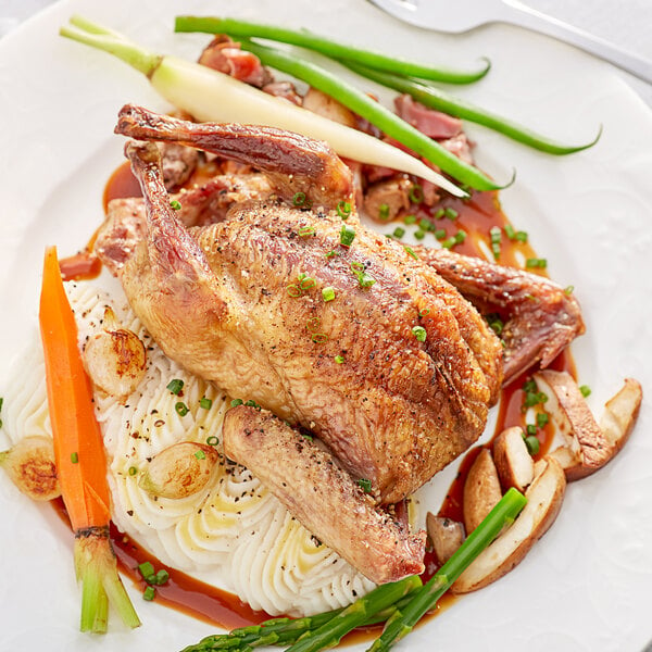 A white plate with a cooked Squab and vegetables with a green leaf.