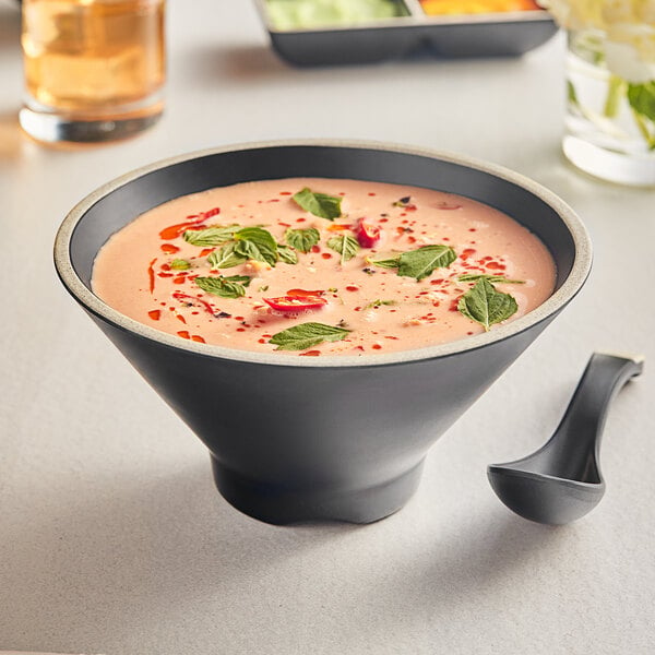 A bowl of soup with an Acopa Ugoki matte black melamine bowl with an ivory rim and a spoon on a table.