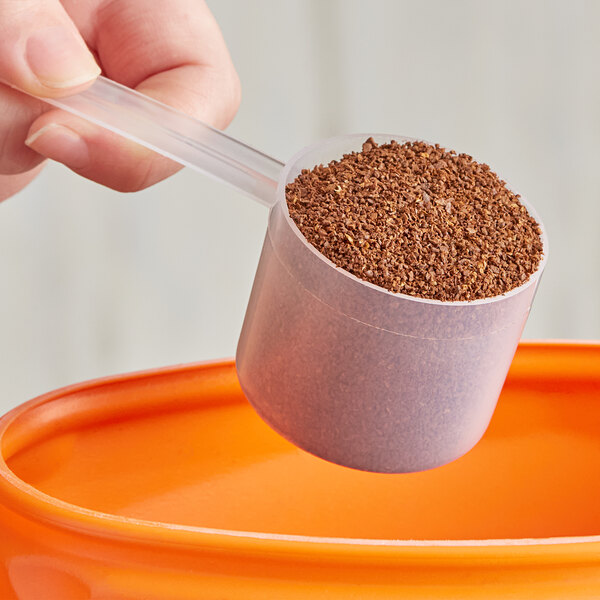 A hand using a 60 cc polypropylene scoop to pour brown powder from a measuring cup into a container.