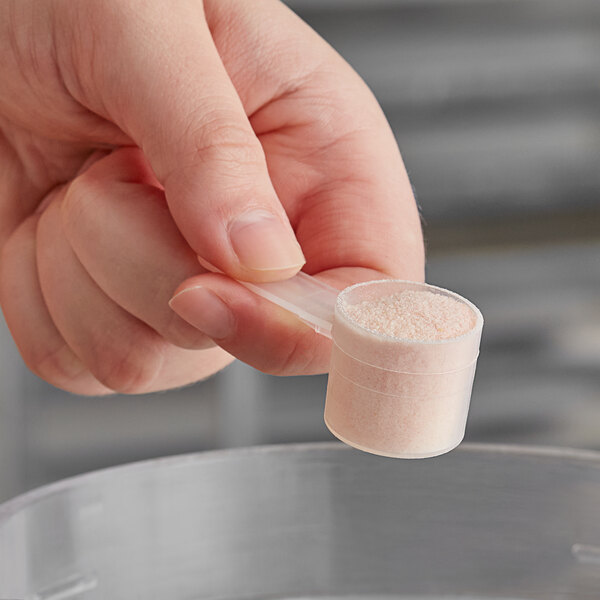 A hand holding a plastic container with a scoop of powder using a 7.5 cc polypropylene scoop.