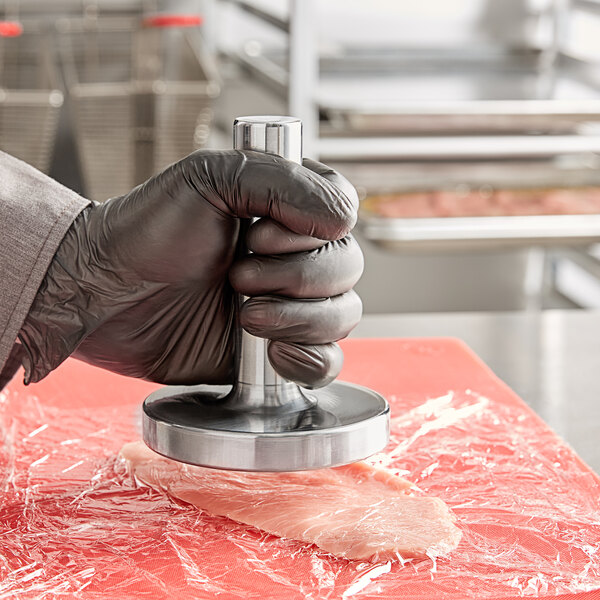 A person in black gloves using an Omcan stainless steel meat tenderizer on a piece of meat on a counter.