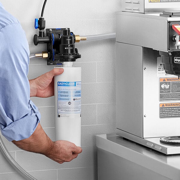 A man holding a Bunn water filter in a white plastic container in a professional kitchen.