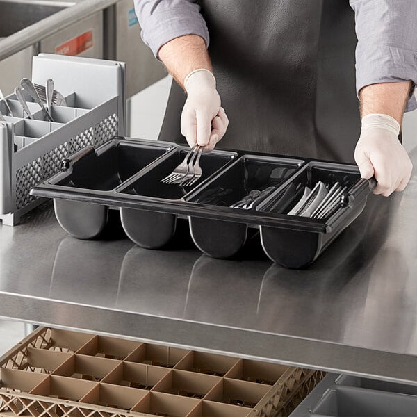 A person in a black apron holding a Choice black plastic cutlery box filled with utensils.