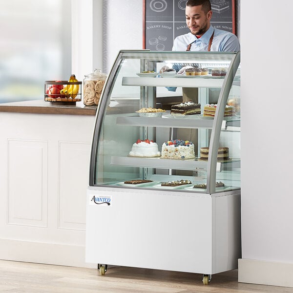 An Avantco refrigerated bakery display case with cakes on shelves behind a man wearing a blue shirt and brown suspenders.