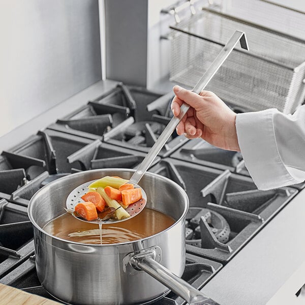 A hand using a Choice stainless steel round perforated skimmer to stir soup in a pot.