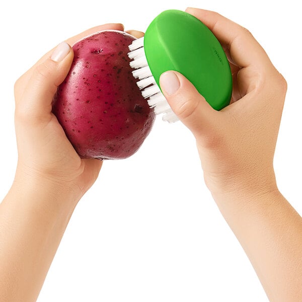 A hand holding a green OXO Good Grips vegetable brush cleaning a potato.
