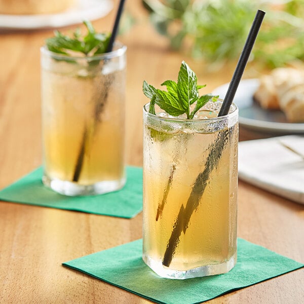 Two glasses of Tractor Beverage Co. green tea with black straws and leaves on a table.